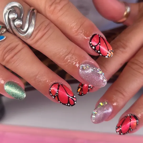 Short almond nails with hand-painted red butterfly wings, a green glitter nail and pink glitter nails topped with crystals