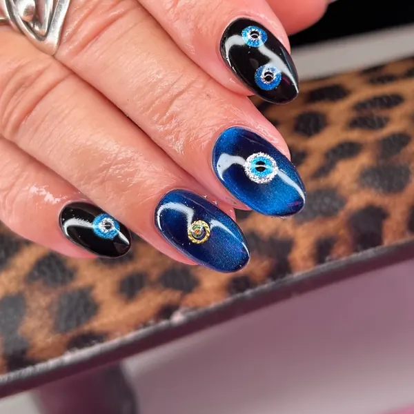 Short almond nails in black and deep blue cat-eye gel with blue evil eye designs and a gold charm