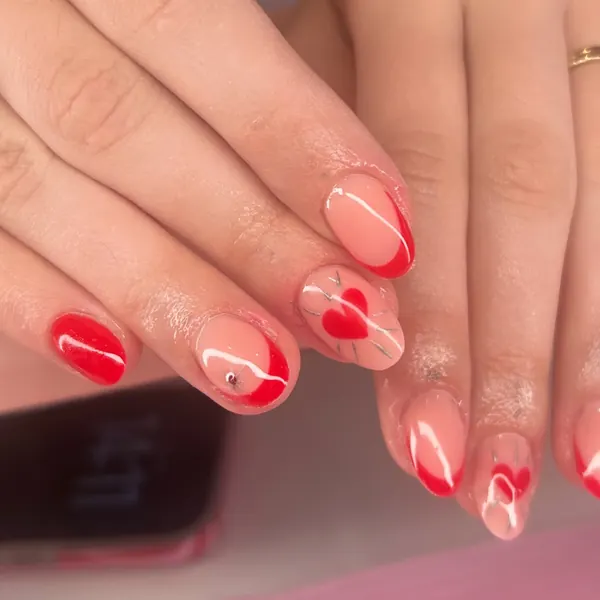 Short almond nails on a nude base with red French tips, hand-painted red hearts, white swirls and a tiny gem, plus one solid red accent nail